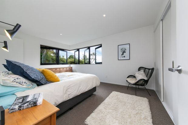 A bright, contemporary bedroom with a large bed, white bedding, colourful accent cushions, a timber bedside table, woven black lounge chair, soft rug, and low-profile built-in storage beneath a corner arrangement of black-framed windows. White walls and ceiling create a clean, airy setting with views of greenery outside.