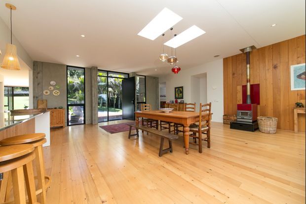 A warm, open-plan dining and living space with extensive light timber flooring and a timber-clad feature wall. The room contains a large traditional timber dining table with ladder-back chairs and a bench, a freestanding red wood-burning stove, exposed timber cabinetry, pendant and recessed lighting, and full-height black-framed glazed doors opening to the garden. A kitchen or bar counter with timber stools is visible at left.