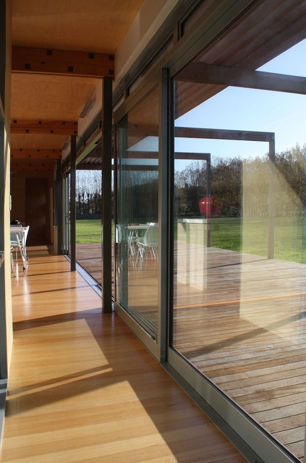 A long, sunlit glazed corridor or indoor-outdoor transition space with extensive floor-to-ceiling sliding glass doors. Warm timber flooring and a timber-lined ceiling continue onto a generous timber deck, with exposed dark timber framing and a lawned garden visible beyond. The image emphasises strong indoor-outdoor connection, reflections, natural light, and clean linear architectural detailing.