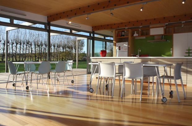 A bright, open-plan contemporary kitchen and dining area with extensive floor-to-ceiling glazing overlooking a tree-lined landscape. The room features a long white kitchen island with integrated seating, a vivid green splashback, white cabinetry, open timber shelving, and multiple white dining tables and wheeled chairs. Warm timber flooring and ceiling, exposed dark structural beams, and strong afternoon sunlight create a spacious, airy atmosphere.