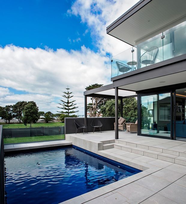 Contemporary multi-level home exterior with a rectangular swimming pool, broad tiled terrace, covered outdoor seating area, frameless glass balcony balustrades, and large glazed sliding doors. The residence has clean horizontal lines, dark trim, pale cladding or rendered surfaces, and a flat roof, set beside a landscaped open area under a bright sky.