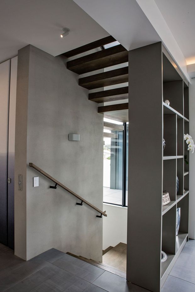 A contemporary interior stairway with dark timber open-riser treads and a matching handrail set against smooth grey plaster walls. A tall grey built-in open shelving partition lines the right side, while a skylight or glazed roof opening brings natural light into the stairwell. Large-format grey floor tiles and black metal stair hardware complete the minimalist composition.