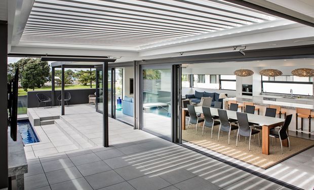 Bright contemporary indoor-outdoor entertaining area with a covered tiled terrace, swimming pool edge, and large black-framed sliding glass openings connecting to an open-plan dining and kitchen space. A linear louvred pergola creates strong striped shadows across the terrace and interior. The kitchen features a long island, white cabinetry, dark benchtops, pendant lights, and a large dining table.