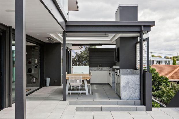 A contemporary rooftop or terrace entertaining area with a covered outdoor kitchen and dining zone. The space features dark grey structural posts and cladding, large light-grey tiled flooring, a marble-look island or benchtop, integrated cabinetry, a dining table with chairs, and a louvred or partially enclosed roof structure. Suburban rooftops and greenery are visible beyond the terrace.