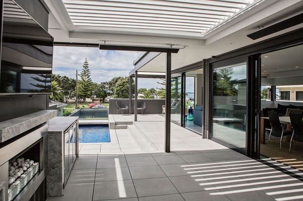 Contemporary covered outdoor entertaining area adjoining a modern home, with large-format pale tiled flooring, a linear pergola or louvred roof casting striped shadows, dark-framed sliding glass doors, a built-in outdoor fireplace and cabinetry, and a swimming pool visible beyond. A dining area is visible through the glazing.