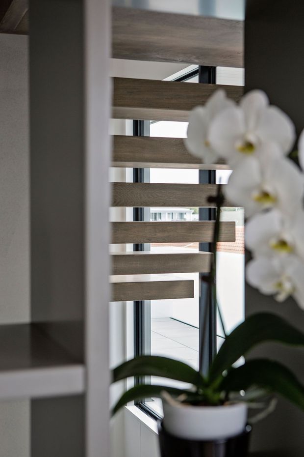 Close-up interior view of a contemporary stair or architectural divider featuring staggered horizontal timber slats mounted between dark metal supports. Large windows and glazing beyond provide natural light, while a white orchid in a planter occupies the foreground.