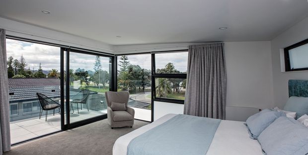 Bright contemporary bedroom with a large bed, upholstered armchair, grey carpet, and full-height corner glazing opening to a balcony or terrace. The room has white walls and ceiling, soft grey curtains, and expansive views of landscaped grounds and neighbouring rooftops.