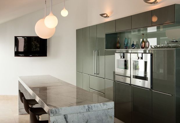 Contemporary kitchen with a large marble island, dark grey-green high-gloss cabinetry, integrated stainless-steel appliances, glass-front display cabinetry, and sculptural pendant lighting. A wall-mounted television is visible on the adjacent wall.