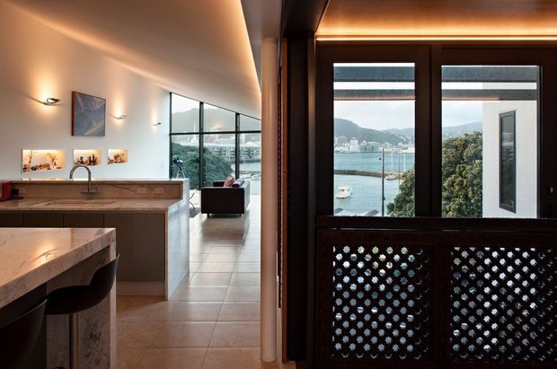 Interior view of a refined open-plan kitchen and living area, with a stone island and timber cabinetry in the foreground, tiled flooring, feature artwork and wall lighting, and a dark timber-framed opening with decorative lattice panels. Large glazing reveals a harbour, boats and surrounding hillside beyond.