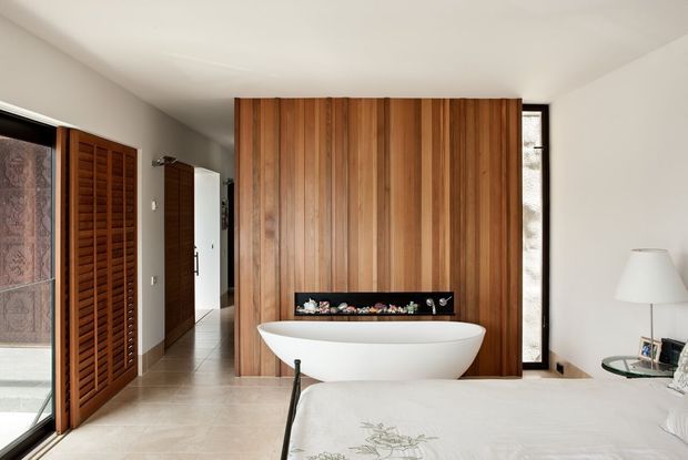 A bright, refined bedroom with a freestanding white bathtub positioned in front of a full-height vertical timber feature wall. Louvered timber doors open toward a bright exterior or adjoining space, while a narrow full-height window, neutral walls, pale flooring, a bedside table, and a simple floor lamp complete the room.