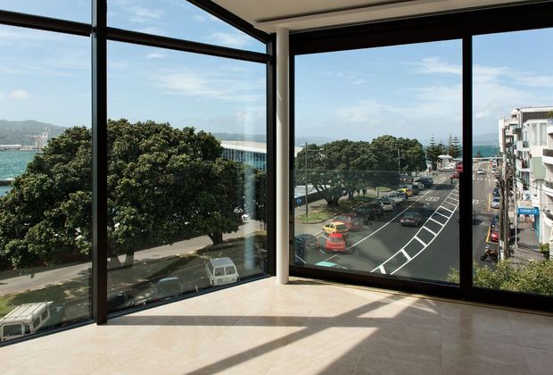 Bright, mostly empty interior corner with full-height black-framed glazing overlooking a coastal urban streetscape, waterfront, trees, and low-rise buildings. Sunlight casts strong geometric shadows across the pale tiled floor; the space appears to be an upper-level commercial or residential room.