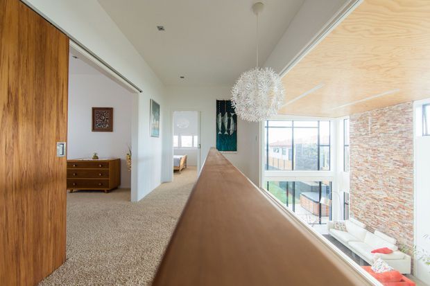 Bright contemporary interior corridor or landing with a long timber built-in balustrade or divider, large windows, exposed brick feature wall, and a sculptural white pendant light. A carpeted bedroom is visible through the open doorway, with timber cabinetry and mid-century-style furnishings. The space overlooks a lower-level living area through expansive glazing.