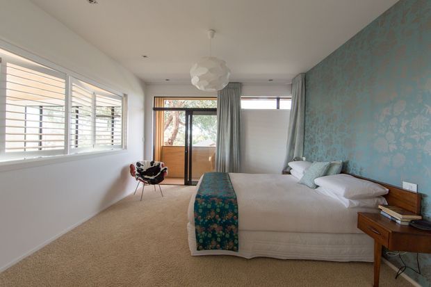 Bright bedroom with cream carpet, white bedding, pale walls, louvered windows, sliding glass doors, and a striking turquoise floral feature wall. A sculptural pendant light, patterned bed runner, upholstered chair, and timber bedside table add decorative character.