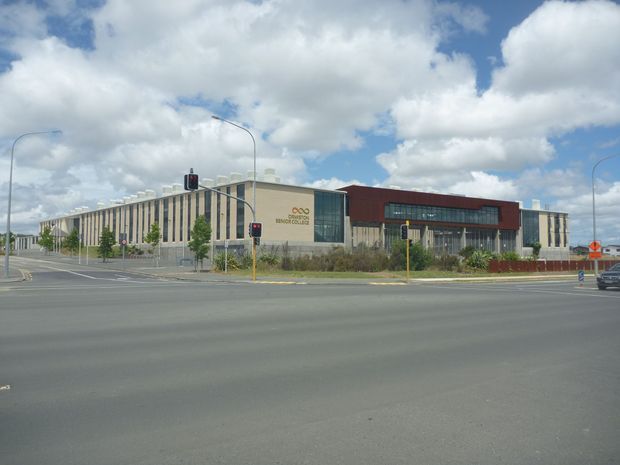 Street-level exterior view of a large contemporary civic or commercial building at a signalised intersection. The building has a long low-rise form, a pale vertical-panel facade, a central glazed entrance, and a prominent dark red-brown upper volume with signage. Broad paved roads and traffic infrastructure occupy the foreground beneath a partly cloudy sky.
