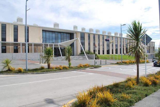Exterior view of a large contemporary institutional or commercial building with a long cream-coloured facade, tall vertical window bays, prominent central curtain glazing, repeated rooftop chimney-like structures, and a projecting angular entrance canopy. The building fronts a landscaped streetscape with low ornamental planting, palms, paved pedestrian areas, and modern street lighting.