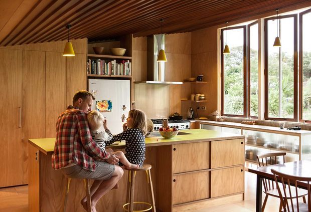 Warm, wood-lined family kitchen with extensive built-in timber cabinetry, a central island with a pale benchtop, open shelving, pendant lighting, and large windows overlooking greenery. A family is gathered at the island, while a dining table is visible beside the windows.