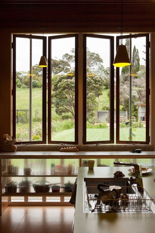 Warm, moody kitchen interior with a long island and integrated sink in the foreground, dark timber-framed awning windows opening to a leafy garden, and suspended cone pendant lights. The room features a timber slatted ceiling, dark timber window frames, reflective patterned cabinetry or backsplash beneath the windows, and a polished warm-toned floor. The composition emphasises the connection between the kitchen and the landscaped outdoors.