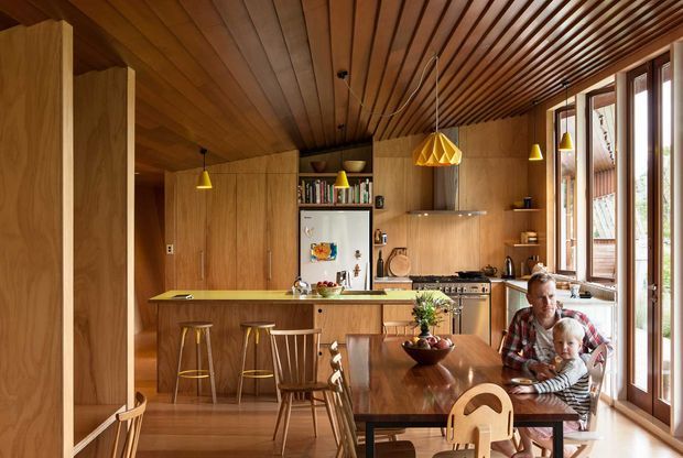 Warm open-plan kitchen and dining area with extensive timber cabinetry, a timber slatted ceiling, island breakfast bar, pendant lighting, stainless-steel appliances, and a wood dining table beside tall glazed doors and windows. A parent and child are seated at the dining table.