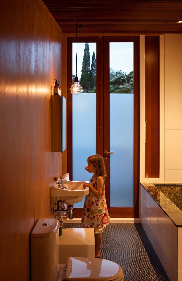 A warmly lit, narrow bathroom with a timber-lined wall and dark timber slatted ceiling. A wall-mounted double basin sits beneath a mirrored cabinet, with a toilet in the foreground and a long stone vanity or counter along the right side. Full-height timber-framed doors with frosted lower glazing bring in filtered daylight and garden views. A child is present near the basin, while the room's fixed finishes and fixtures dominate the image.