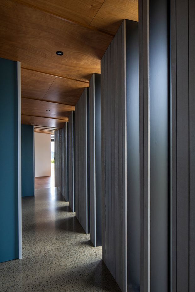 A long, narrow modern hallway with a warm timber-lined ceiling, dark vertically ribbed wall panels and recessed openings along one side, deep blue painted walls, and a polished terrazzo-like speckled floor. Recessed ceiling lights and daylight at the far end emphasize the corridor’s strong linear perspective and repeating architectural rhythm.