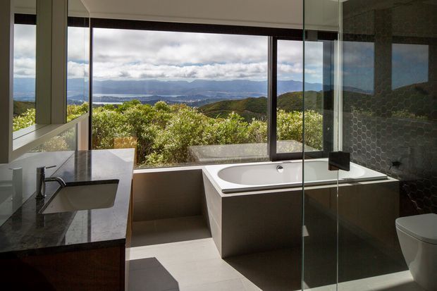 Contemporary bathroom with a freestanding white bathtub positioned beside expansive floor-to-ceiling glazing, overlooking a lush hilly landscape and distant water. The room includes a dark stone vanity benchtop with an integrated rectangular basin, dark timber cabinetry, a frameless glass shower enclosure, dark hexagonal wall tiles, and a white toilet. Natural light and the panoramic outlook create a refined, serene atmosphere.