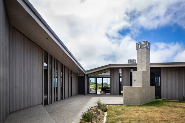 Exterior view of a contemporary single-storey courtyard house arranged around a paved central courtyard. The building has dark vertical wall cladding, extensive floor-to-ceiling glazing, low-pitched roof planes, and a prominent pale masonry chimney with a louvered cap. Sparse landscaping and a partly cloudy sky are visible.