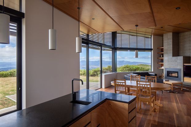 Open-plan kitchen and dining/living area with extensive floor-to-ceiling glazing overlooking a hilly landscape. The kitchen features a dark stone island benchtop, timber cabinetry, a black sink tap and integrated appliances. A timber-lined ceiling, pendant lights, timber flooring, built-in shelving and a tiled fireplace wall create a warm contemporary interior.