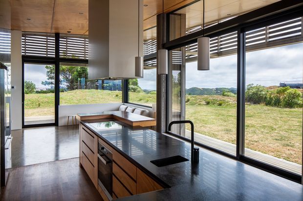A contemporary open-plan kitchen and dining area with a large timber island, dark stone benchtop, integrated sink, pendant lights, extensive black-framed glazing, and horizontal external sun louvres. The room opens toward a grassy rural landscape with hills beyond, while a built-in dining nook sits beside the windows. Timber-lined ceilings and polished concrete flooring create a warm modern interior.