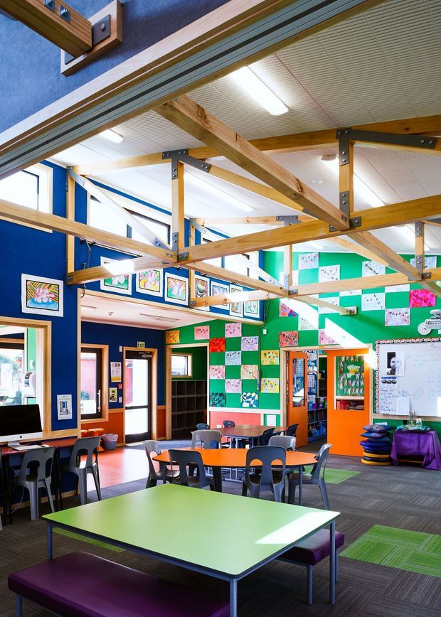 A bright, colourful school or community learning space with multiple green and orange tables, black chairs, upholstered benches, a computer workstation, bookshelves, a whiteboard and children's artwork displayed across vivid blue and green walls. Exposed timber post-and-beam framing with dark metal connectors spans the high ceiling, which includes white acoustic panels and fluorescent strip lighting. The room has carpet tile flooring and extensive glazing, creating an energetic, playful educational interior.