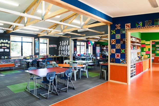 Bright, colourful school or educational common room with grouped tables and chairs, display shelving, craftwork and noticeboards. The open-plan interior has exposed timber roof trusses, dark blue walls, vibrant green and orange adjoining areas, and patterned carpet tiles transitioning to orange resilient flooring.