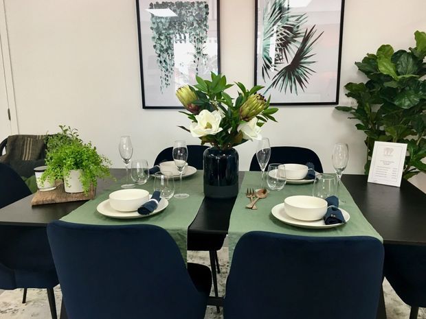 A refined dining area with a dark rectangular dining table set for four, featuring white crockery, wine glasses, cutlery, and soft green table runners. Deep navy upholstered dining chairs surround the table, while a black vase of white and green flowers forms the centrepiece. Large botanical prints, indoor plants, and neutral walls create a fresh, elegant atmosphere.