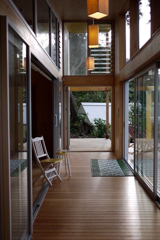 A narrow, light-filled timber-lined passage or glazed entry gallery with warm wood flooring, extensive floor-to-ceiling glazing, sliding glass doors, and a tall clerestory-like volume. Three suspended timber pendant lights hang above the corridor, while a simple folding chair and small side table sit beside the entrance. The view opens toward a covered exterior deck and dense greenery, creating a strong indoor-outdoor connection.