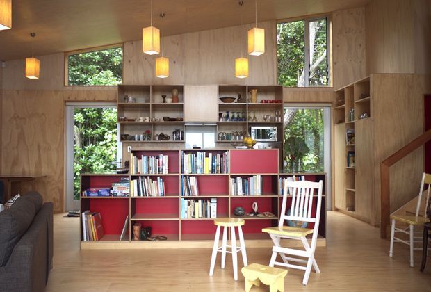 A warm, timber-lined interior centred on a large built-in red and natural-wood shelving and display unit filled with books and objects. Floor-to-ceiling and high-set windows look onto leafy greenery, while several simple pendant lights hang from the timber ceiling. A sofa is partially visible at left, with white folding chairs and a small stool in the foreground; a timber stair and additional shelving appear at right.