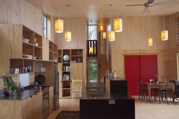 Open-plan kitchen and dining area in a timber-lined interior, featuring extensive plywood cabinetry and shelving, a dark central island, warm pendant lighting, a dining table, and a staircase leading to an upper level. The space has a warm, natural, contemporary character with visible timber wall and ceiling finishes.