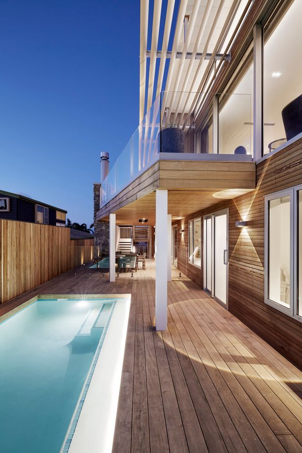 A contemporary multi-level timber-clad home with an elevated balcony and pergola-like overhead slats overlooking a narrow swimming pool. The outdoor deck extends along the house, with glazed balustrades, white structural posts, large windows and sliding doors, warm timber wall cladding, and an outdoor dining area beneath the upper level.