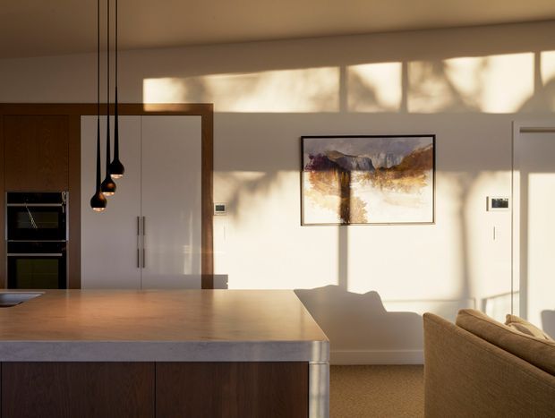 Warm, sunlit open-plan kitchen and living area. A large timber island with a pale stone benchtop dominates the foreground, with dark timber cabinetry, integrated appliances, tall white pantry cabinetry, and three black pendant lights. A neutral upholstered sofa, framed artwork, and strong afternoon shadows create a calm contemporary interior.