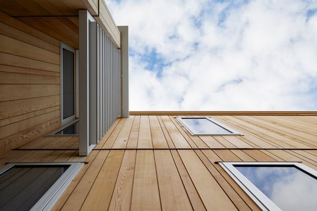 Upward-looking close view of a modern building facade clad in warm vertical timber boards, with white-framed windows, a projecting screened balcony or louvred element, and a flat roof edge against the sky.