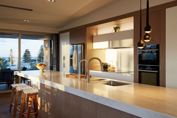 Contemporary kitchen with a large light stone island and integrated sink, timber-look cabinetry, dark built-in ovens, warm recessed and pendant lighting, and several timber bar stools. Full-height glazing beside the kitchen opens views toward trees and water, creating a bright indoor-outdoor connection.
