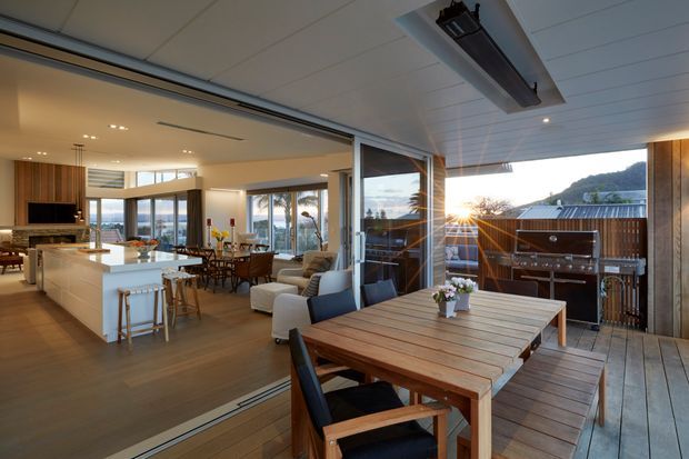 Open-plan contemporary home interior extending onto a timber deck, with a white kitchen island, dining and living areas, extensive glazing, and an outdoor dining setting beside a built-in barbecue. Warm sunset light and timber finishes create a relaxed indoor–outdoor atmosphere.