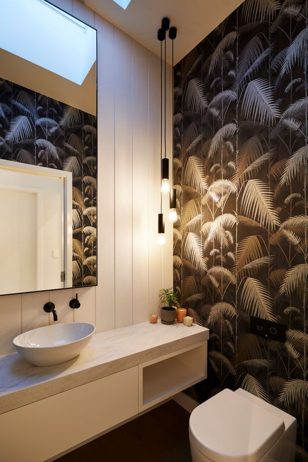 A stylish contemporary bathroom featuring a floating pale vanity with an integrated stone benchtop, white vessel basin, black wall-mounted tapware, and a modern white toilet. One wall is covered in dramatic dark tropical palm-leaf wallpaper, contrasted by light vertical wall panelling and a large mirror. Three suspended black pendant lights provide warm illumination, with a skylight or ceiling opening visible above.