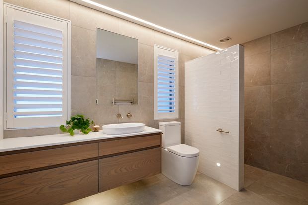 Contemporary bathroom with a floating timber vanity, white vessel basin, wall-mounted mirror, toilet, and a glass-enclosed shower area behind a partial white tiled partition. Large beige stone-look tiles cover the walls and floor, with white plantation shutters and integrated linear ceiling lighting.