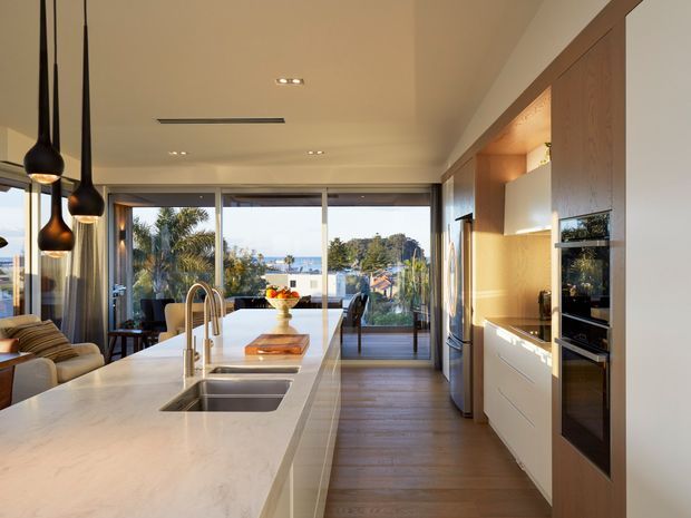 A contemporary open-plan kitchen viewed along a long white stone island toward a glazed dining and living area. The kitchen features integrated timber cabinetry, white drawers, stainless-steel appliances, a double-bowl sink with brushed metal tapware, dark timber flooring, and sculptural black pendant lights. Floor-to-ceiling glazing opens the room to a leafy suburban outlook and balcony.