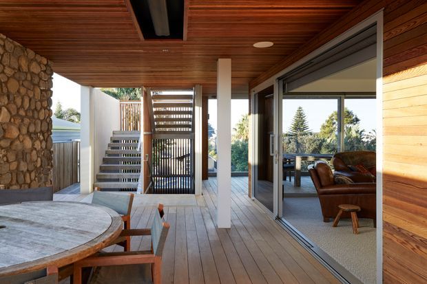 Covered timber-lined outdoor living and dining area adjoining a glazed interior lounge. The space features wide timber decking, a round outdoor dining table, a stone-clad wall, exposed timber ceiling, large sliding glass doors, and a stairway leading to an upper level. A leather lounge and garden views are visible through the glazing.