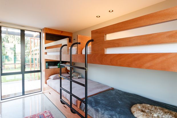 Compact contemporary bedroom with built-in timber bunk beds and integrated shelving, black metal ladder rails, pale walls, polished light flooring, and large glazed sliding doors opening to an outdoor area. The room has warm timber detailing and a practical space-saving layout.