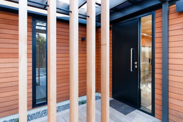 Contemporary residential entry facade featuring warm horizontal timber cladding, a dramatic matte-black pivot door with a tall vertical handle, narrow glazed sidelights, pale timber vertical screening battens, and dark metal framing. A partially covered entry walkway is finished with large light-grey pavers and decorative white stone landscaping, creating a crisp contrast between natural timber and black metal.