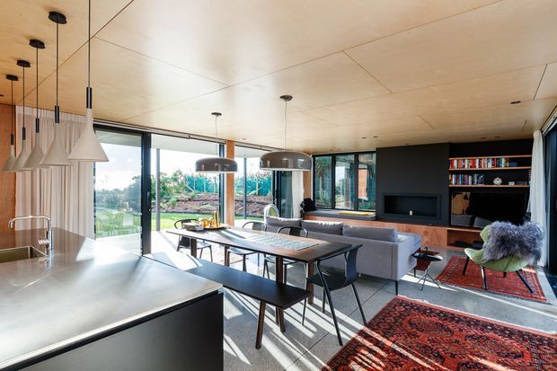 Open-plan contemporary living and dining area viewed from the kitchen, with a large island in the foreground, timber-lined ceiling, extensive black-framed glazing, a dining table with bench seating, and a lounge with built-in dark timber shelving and television cabinetry. Pendant lights, layered rugs, and warm timber finishes create a refined, inviting interior with strong indoor-outdoor connection.