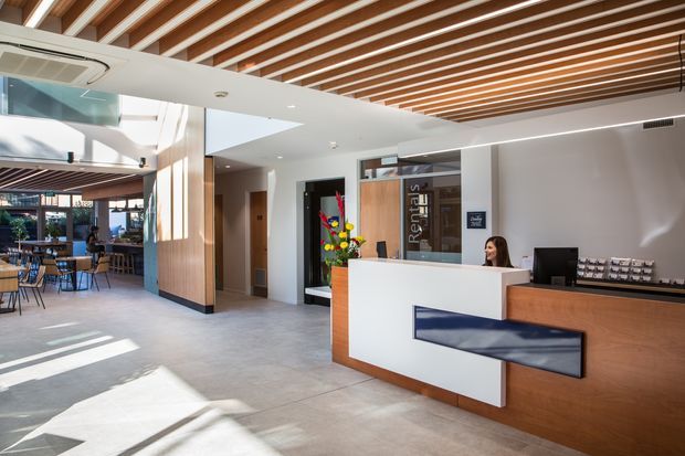 A bright contemporary reception lobby with a white and timber reception desk, integrated blue accent panel, and a staff workstation. The space features a pale tiled floor, white walls, extensive glazing, and a ceiling of exposed timber battens with recessed lighting. A furnished café or lounge area is visible beyond the reception zone.