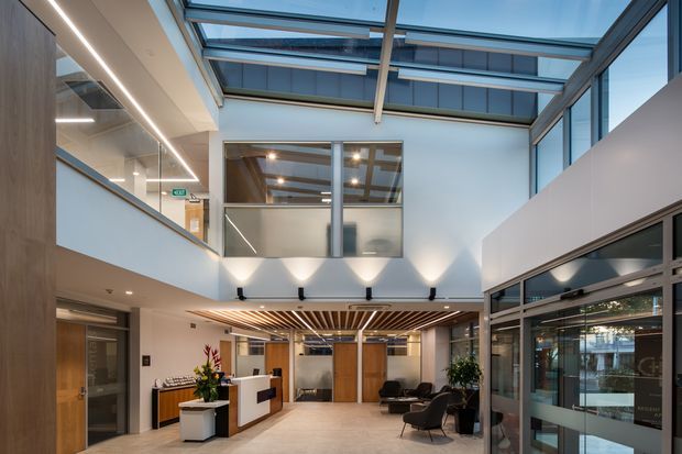 A bright contemporary commercial interior, likely an office or reception lobby, viewed from an open ground-floor area beneath a glazed atrium. The space features a double-height void, extensive clerestory and roof glazing, timber wall panelling, a reception desk, lounge seating, glass partitions, linear recessed lighting, and a timber-slat ceiling feature. The palette is predominantly white, light timber, grey, and black, creating a refined and airy atmosphere.