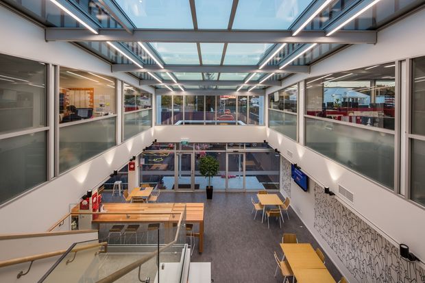 Bright contemporary commercial interior viewed from an upper landing, overlooking a multi-level atrium or circulation space. The design features extensive internal glazing, exposed structural framing, linear LED lighting, skylight-like glazed roof panels, staircases with glass balustrades, timber café-style tables, and a patterned feature wall. The space appears open, functional, and brightly lit.
