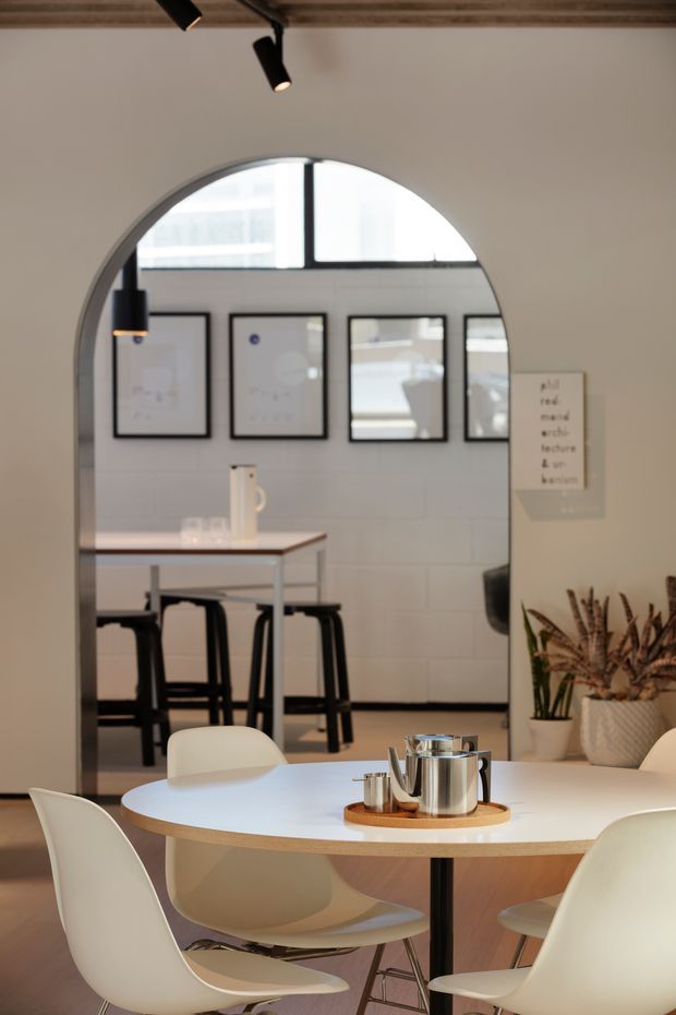 A softly lit contemporary dining and meeting area viewed through a large arched opening. A round white table with a pale timber edge and sculptural white chairs occupies the foreground, with a stainless-steel tea service on a tray. Behind it is a high table with black stools, framed artwork, tiled walls, and large internal glazing. The space has a restrained neutral palette, black accents, indoor plants, track lighting, and a timber-lined ceiling.